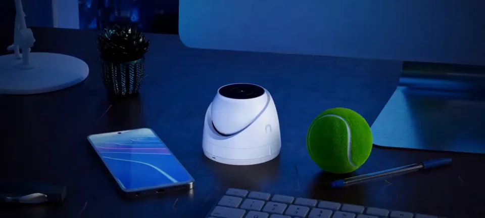 White UniFi G5 Turret Ultra sitting on a desk next to a phone and a tennis ball, showing its compact size