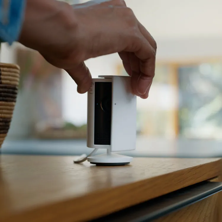 A hand sliding the detachable lens cover across a Ring Indoor Cam Plus on a wooden table