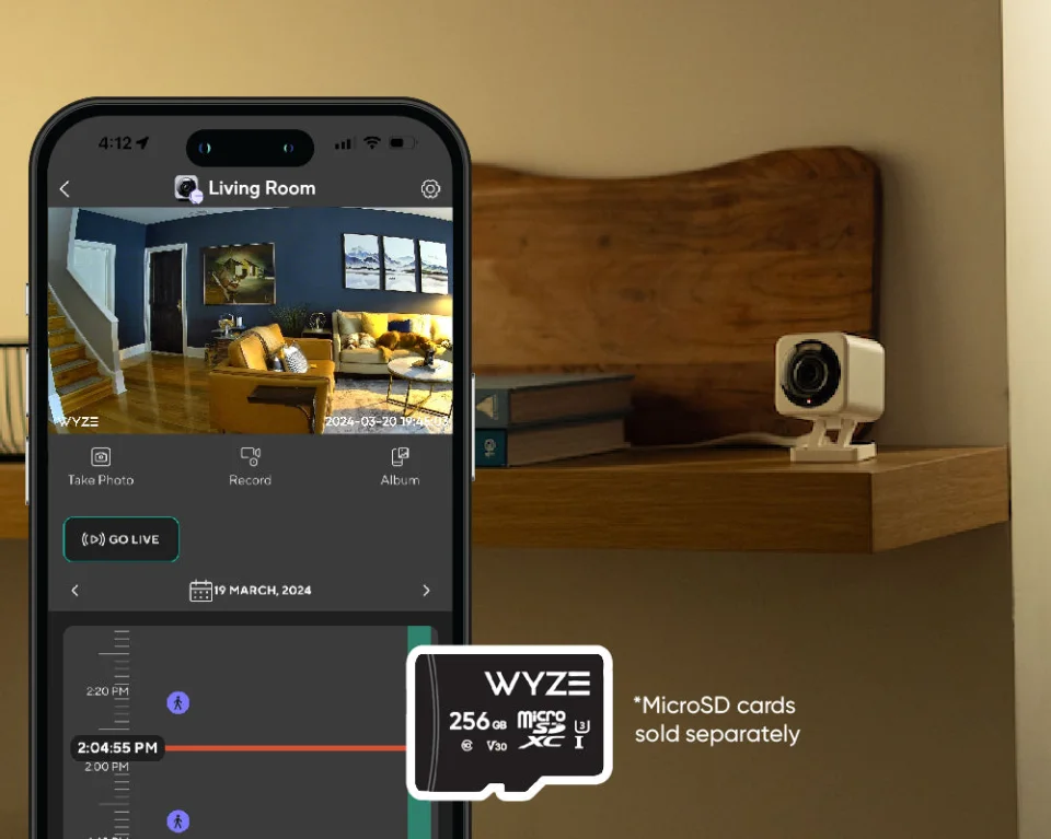 Wyze Cam v4 on a shelf beside a phone showing the Wyze app timeline and a Wyze microSD card