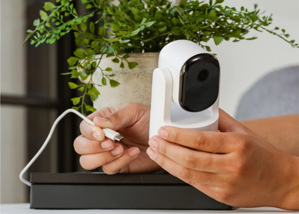 Hands holding the Arlo Essential Pan Tilt Indoor camera and its USB-C power cable in front of a plant