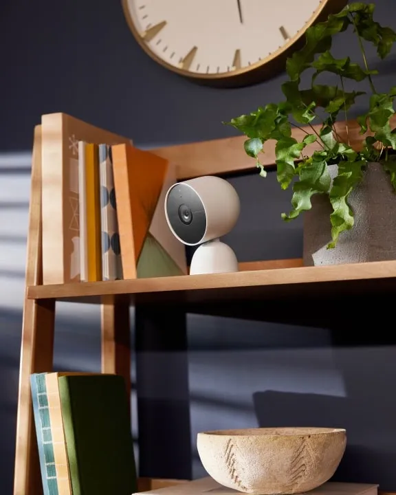 Google Nest Cam Indoor 3rd Gen in Snow sitting on a wooden bookshelf next to books and a plant